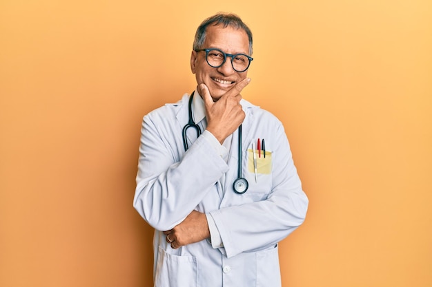 Hombre indio de mediana edad con abrigo médico y estetoscopio mirando confiado a la cámara sonriendo con los brazos cruzados y la mano levantada en la barbilla. pensando en positivo.