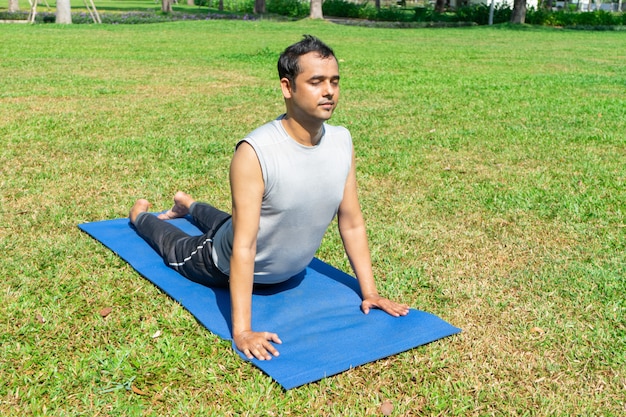 Hombre indio que hace actitud ascendente del perro al aire libre en césped verde. Concepto de yoga al aire libre