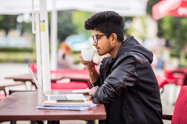 Hombre indio usando laptop mientras bebe una taza de café en un café callejero al aire libre