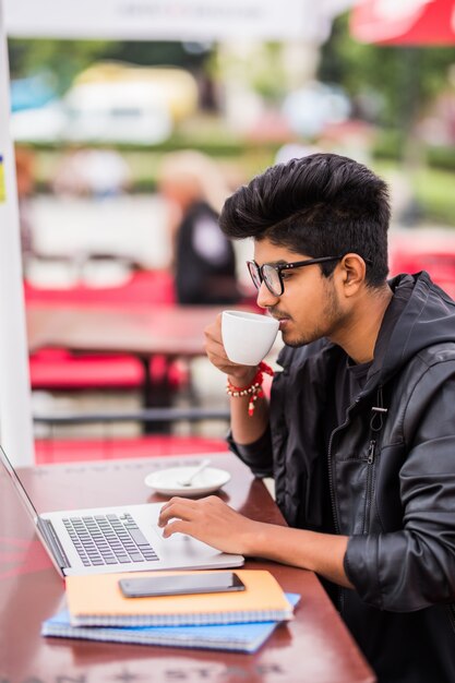 Hombre indio usando laptop mientras bebe una taza de café en un café callejero al aire libre