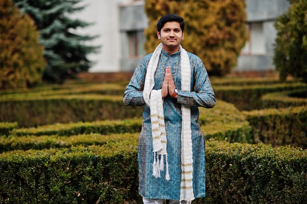 Hombre indio vestido con ropa tradicional con bufanda blanca posada al aire libre contra arbustos verdes en el parque muestra el signo de las manos de namaste