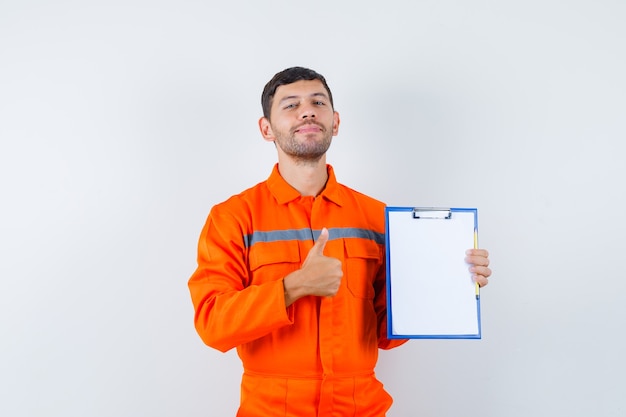 Hombre industrial sosteniendo el portapapeles, mostrando el pulgar hacia arriba en uniforme y mirando contento, vista frontal.