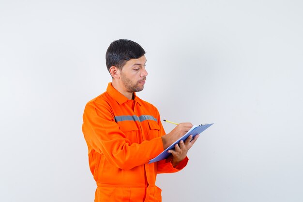 Hombre industrial tomando notas en el portapapeles en uniforme y mirando ocupado, vista frontal.