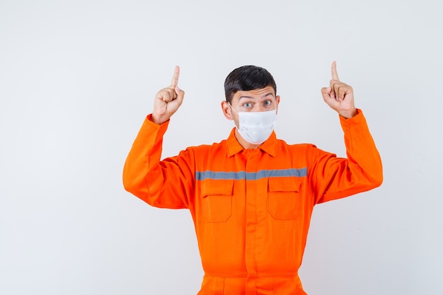 Foto gratuita hombre industrial en uniforme, máscara apuntando con el dedo hacia arriba y mirando curioso, vista frontal.