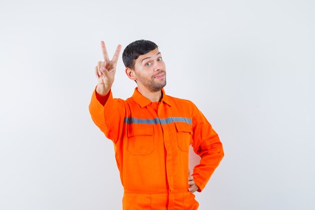 Hombre industrial en uniforme mostrando v-sign y mirando alegre, vista frontal.