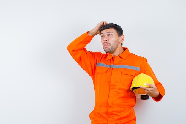 Hombre industrial en uniforme sosteniendo casco, rascándose la cabeza y mirando pensativo, vista frontal.