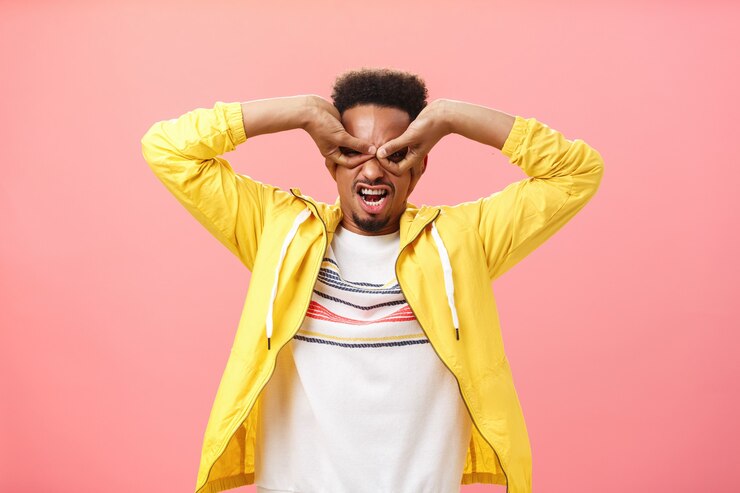 Foto gratuito el hombre desde la infancia soñó convertirse en superhéroe. chico joven afroamericano juguetón infantil actuando inmaduro haciendo máscara de manos sobre los ojos aping de pie en chaqueta amarilla sobre pared rosa