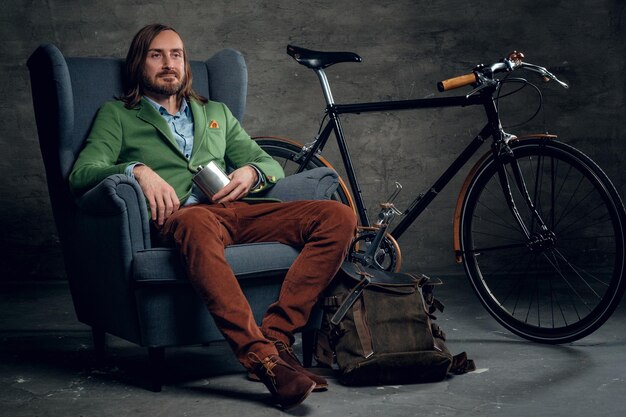 Un hombre informal con el pelo largo y rubio vestido con una chaqueta verde se sienta en una silla con una bicicleta de una sola velocidad en el fondo.