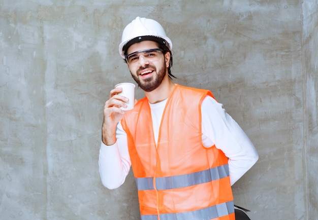 Hombre ingeniero en casco blanco y anteojos protectores sosteniendo una taza de bebida.