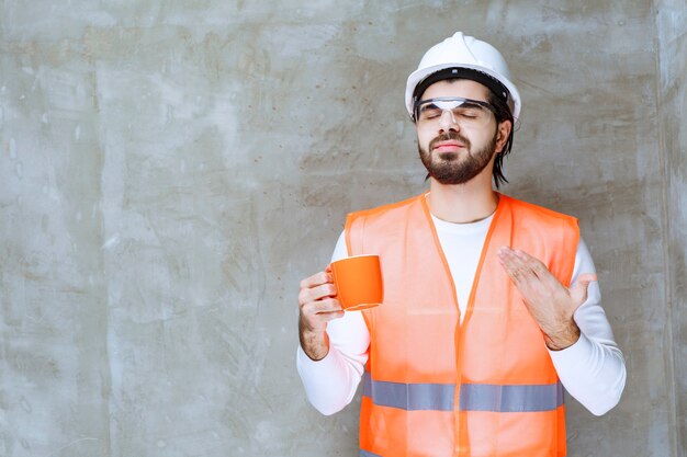 Hombre ingeniero en casco blanco sosteniendo una taza naranja.