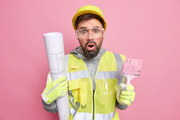 hombre ingeniero posa con el plano de la herramienta de construcción sorprendido de tener mucho trabajo vestido con uniforme de trabajo yendo a pintar paredes en una casa nueva después de la reconstrucción. Mejora de apartamento