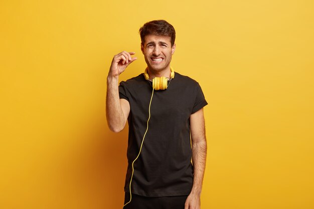 Hombre insatisfecho aprieta los dientes, muestra un pequeño gesto, dice un poco más, usa una camiseta negra informal, tiene auriculares para escuchar la lista de reproducción