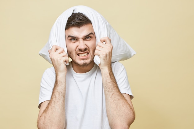 Hombre de insomnio, privación del sueño y concepto de problema de sueño. Joven deprimido con barba cubriendo las orejas y la cabeza, tratando de bloquear un sonido de alarma ruidoso o despertado por la noche por un vecino ruidoso