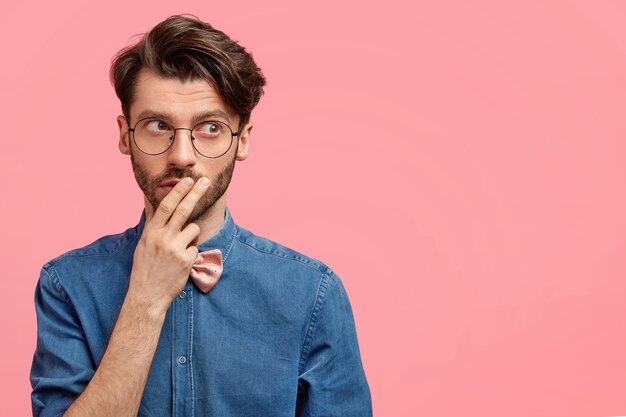 Hombre inteligente sin afeitar reflexivo mira pensativamente a la distancia, toca la boca con la mano, está sumido en sus pensamientos, piensa en algo importante, vestido con una chaqueta de mezclilla, aislado sobre una pared rosa