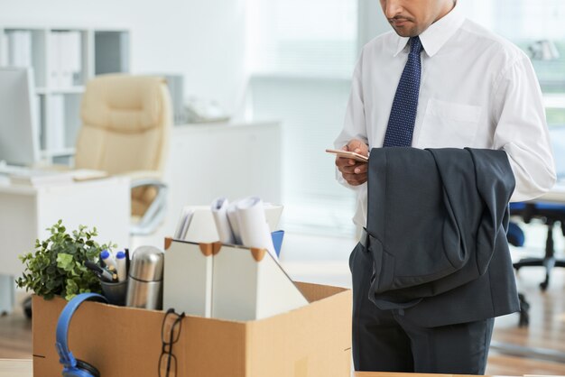 Hombre irreconocible parado en la oficina y usando un teléfono inteligente, con pertenencias personales en caja