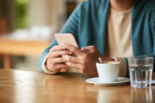 Hombre irreconocible sentado en la cafetería con una taza de café y agua y usando el teléfono inteligente