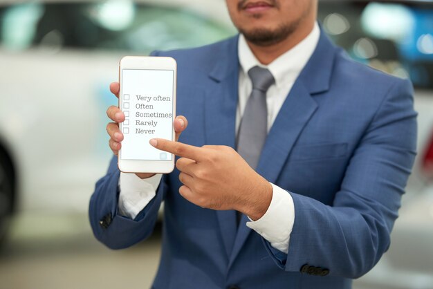 Hombre irreconocible en traje de negocios sosteniendo el teléfono inteligente y apuntando a la encuesta en pantalla