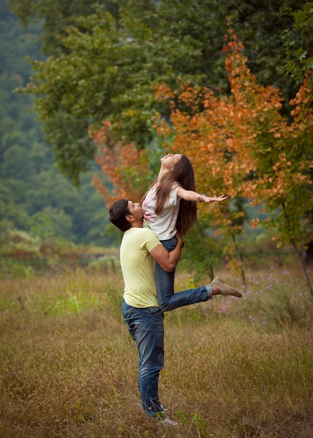 Un hombre en jeans y camisa con una mujer en sus brazos en el bosque y mirando feliz durante el día.