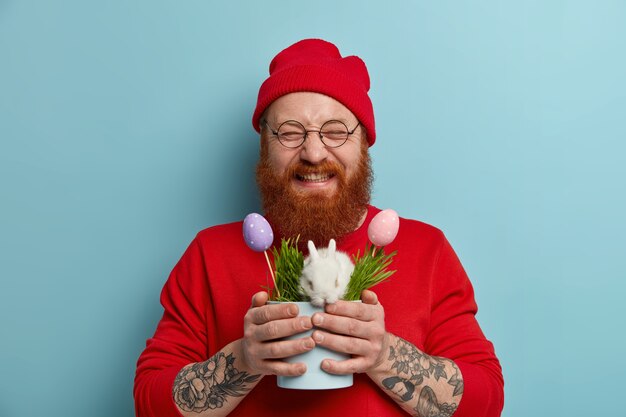 Hombre de jengibre feliz lleno de alegría satisfecho después de la búsqueda de huevos, sostiene una olla con un conejito de Pascua blanco en la hierba y huevos coloridos, viste un traje rojo, gafas redondas, celebra las vacaciones. Concepto de tiempo de primavera