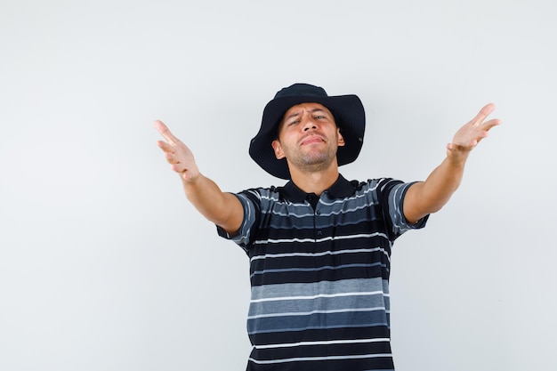 Hombre joven abriendo los brazos para abrazar en camiseta, sombrero y mirando amigable, vista frontal.