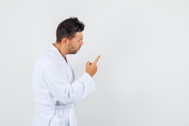 Hombre joven advirtiendo a alguien con gesto de dedo en bata de baño blanca y mirando nervioso, vista frontal.
