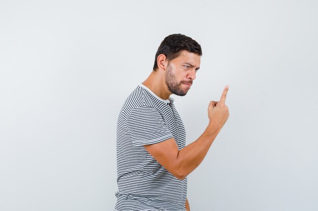 Hombre joven advirtiendo con el dedo en la camiseta y mirando enojado, vista frontal.