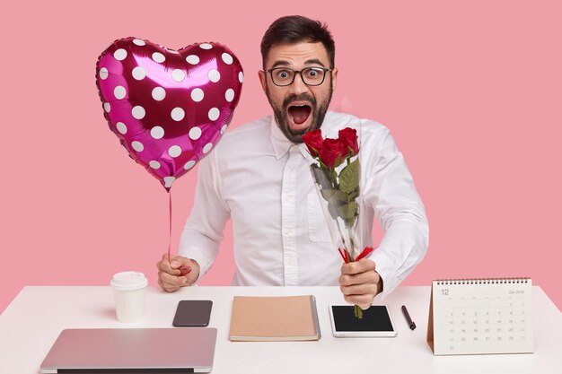 Un hombre joven sin afeitar sorprendido lleva un ramo y un globo en forma de San Valentín, sorprendido al escuchar el cumplido