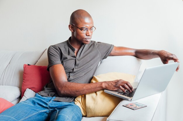 Hombre joven africano serio que se sienta en el sofá usando el ordenador portátil en casa