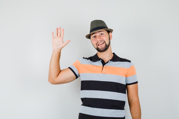 Hombre joven agitando la mano para saludar en camiseta, sombrero y alegre.