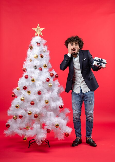 Hombre joven agotado sosteniendo su regalo de pie cerca del árbol de Navidad blanco decorado en el lado derecho del rojo