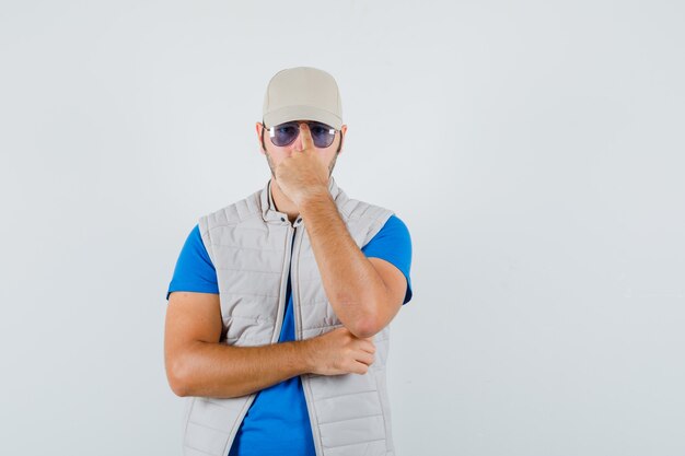 Hombre joven ajustando sus gafas en camiseta, chaqueta, gorra y mirando confiado. vista frontal.