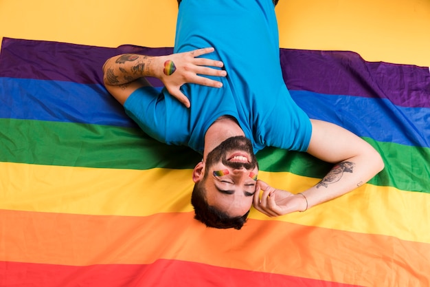 Hombre joven al revés con el arco iris en la cara acostado y sonriendo en la bandera LGBT