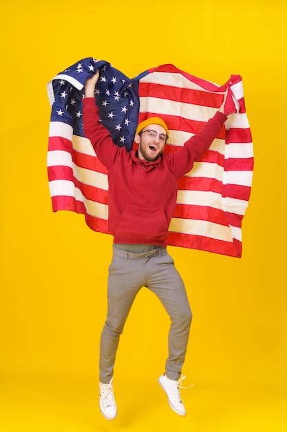 Hombre joven alegre con bandera americana Hombre joven divertido feliz en sudadera con capucha roja y bandera de Estados Unidos saltando