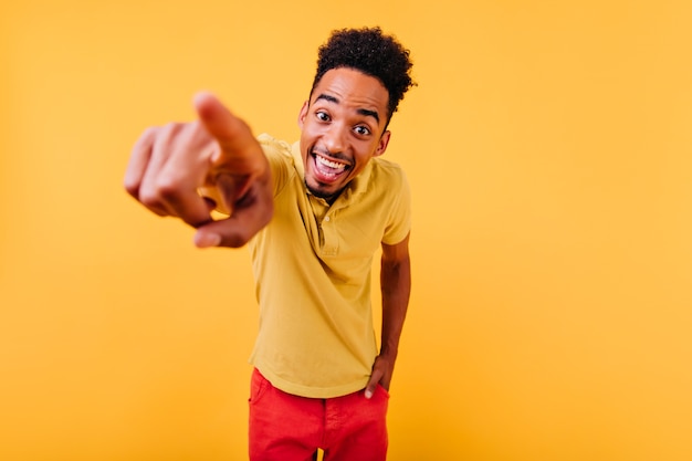 Hombre joven alegre con el dedo acusador de sonrisa sorprendida. Filmación en interiores del encantador niño africano aislado.