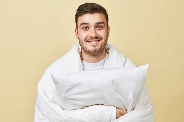 Hombre joven alegre positivo con linda sonrisa y rostro sin afeitar de pie en la pared en blanco, envuelto en una manta blanca, sintiéndose lleno de alegría, recuperándose del frío, sosteniendo la almohada, yendo a dormir en la cama
