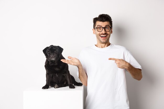 Hombre joven alegre que señala con el dedo a su perro, mostrando pequeño pug negro lindo sentado, fondo blanco.