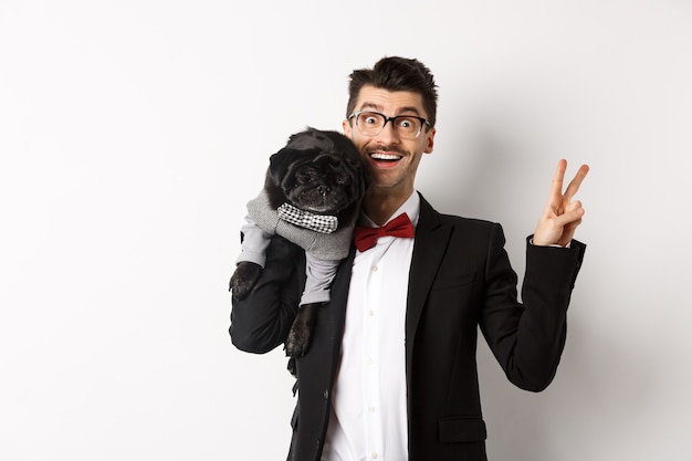 Hombre joven alegre en traje y gafas tomando fotos con lindo perro pug negro en su hombro, sonriendo feliz y mostrando el signo de la paz, posando sobre fondo blanco.