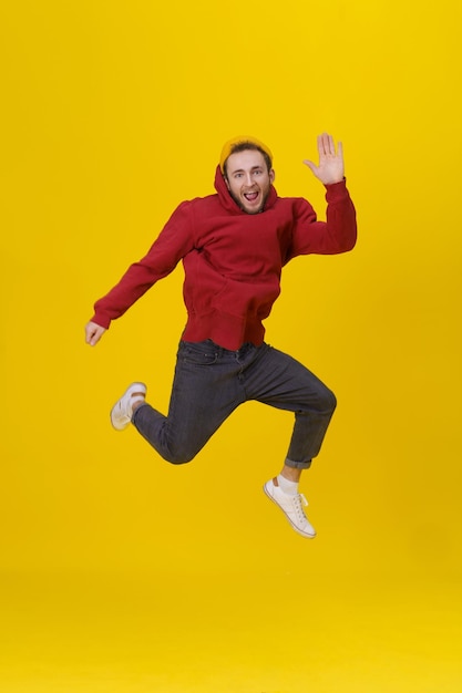 Hombre joven de alegría saltando alto con sudadera con capucha roja casual y jeans aislados en amarillo Chico hipster humorístico en salto
