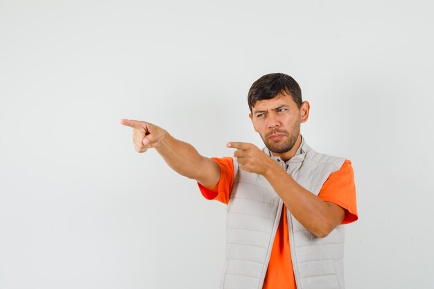 Hombre joven apuntando hacia afuera en camiseta, chaqueta y mirando enfocado, vista frontal.