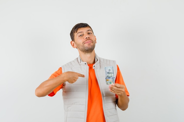 Hombre joven apuntando al billete de un dólar en camiseta, chaqueta y mirando confiado. vista frontal.