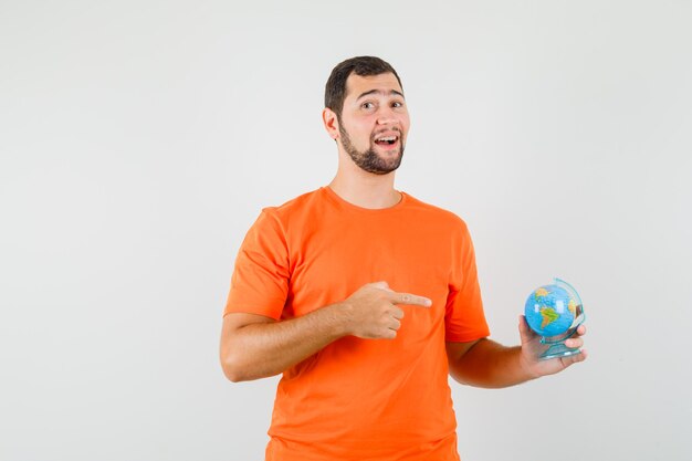 Hombre joven apuntando al globo terráqueo en camiseta naranja y mirando alegre, vista frontal.