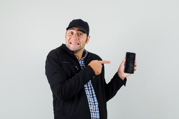 Hombre joven apuntando al teléfono móvil en camisa, chaqueta, gorra y mirando contento.