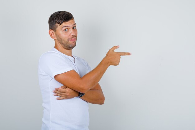 Hombre joven apuntando a algo con el dedo en camiseta blanca y mirando positivo. .