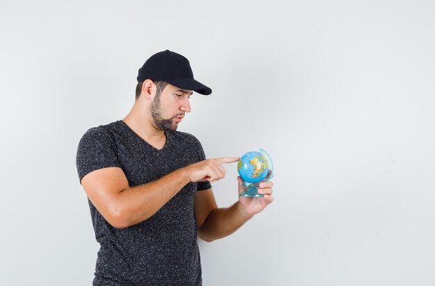 Hombre joven apuntando a algún lugar del globo en camiseta y gorra y mirando pensativo