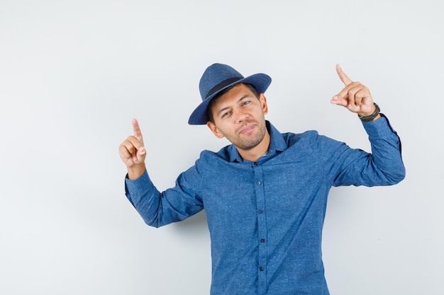 Hombre joven apuntando hacia arriba con camisa azul, sombrero y aspecto fresco. vista frontal.