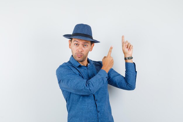 Hombre joven apuntando hacia arriba con camisa azul, sombrero y mirando curioso, vista frontal.
