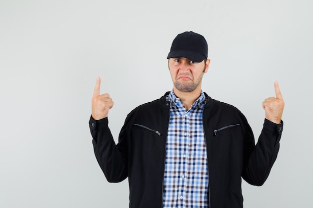 Foto gratuita hombre joven apuntando hacia arriba en camisa, chaqueta, gorra y mirando disgustado, vista frontal.