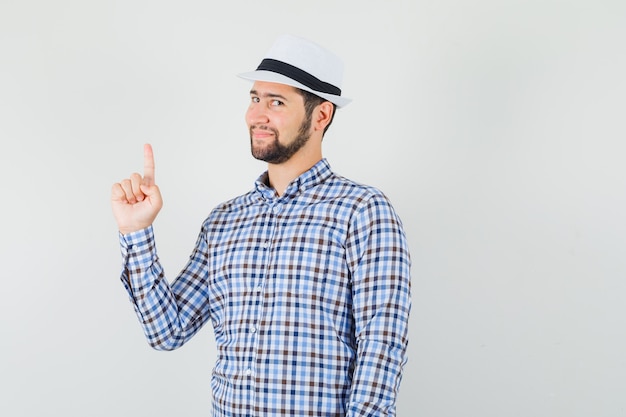 Hombre joven apuntando hacia arriba con camisa a cuadros, sombrero y aspecto sensato.