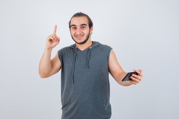 Hombre joven apuntando hacia arriba con camiseta con capucha y mirando feliz, vista frontal.