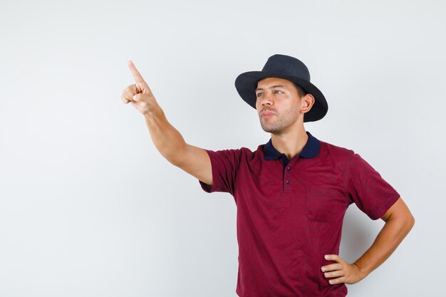 Hombre joven apuntando hacia arriba en camiseta, sombrero y mirando enfocado, vista frontal.